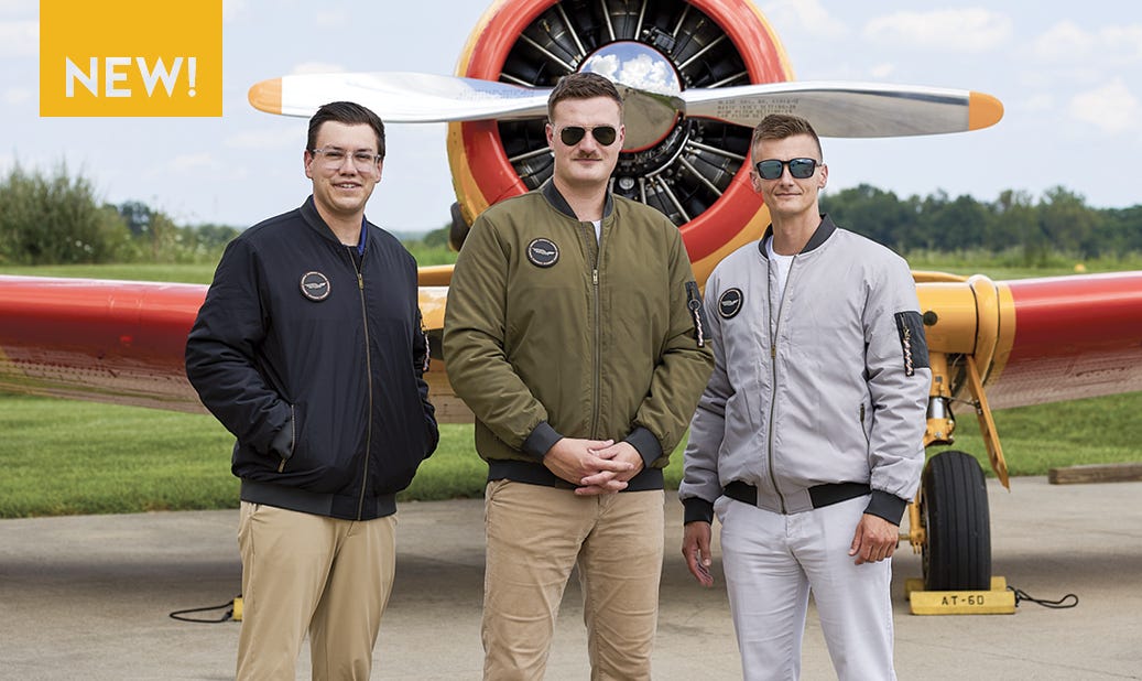 The Modern MA-1 AeroWear™ Jacket. Timeless design with modern versatility-lightweight, durable and built for all-season wear. 3 models wearing blue, green and grey jackets in front of vintage aircraft.