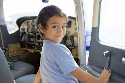 A young child standing outside of a small airplane, smiling back at the camera